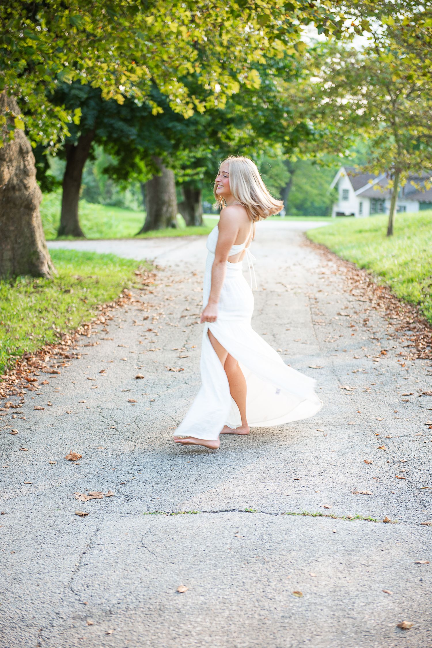A flowing white dress twirls gracefully on a sunlit autumn path lined with golden leaves and trees.