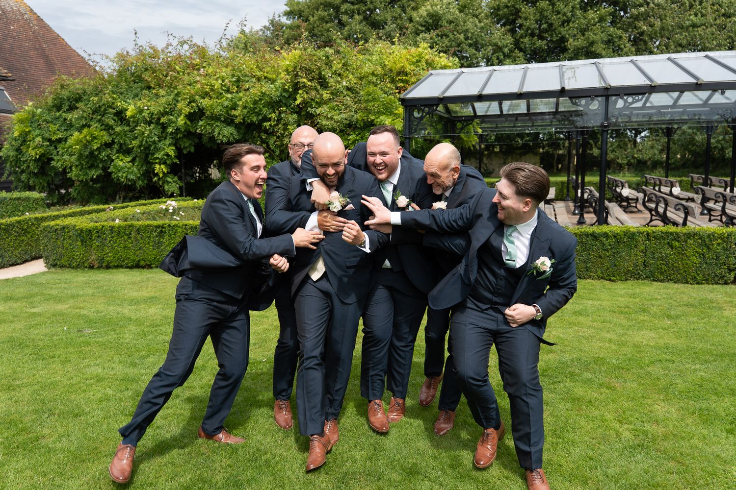 A group of people in formal attire jumping in sync on a green lawn during what appears to be a wedding celebration.
