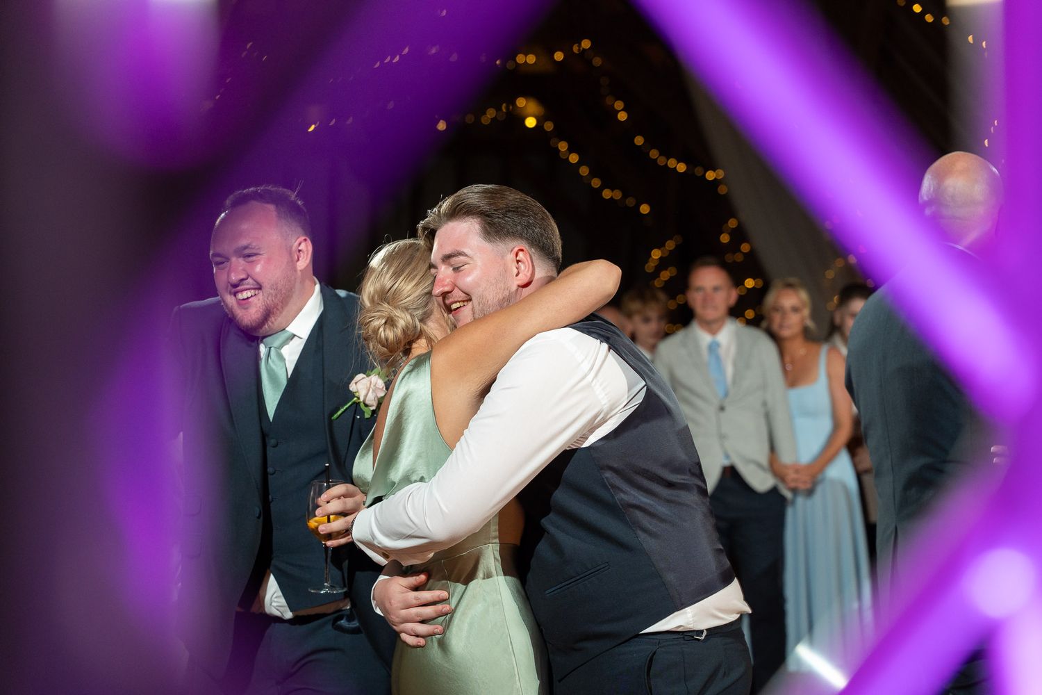 Wedding guests dance and celebrate on a purple-lit dance floor during an evening reception.