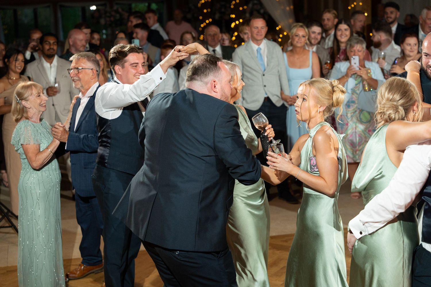 Wedding guests dance and celebrate on a crowded reception dance floor with string lights overhead.