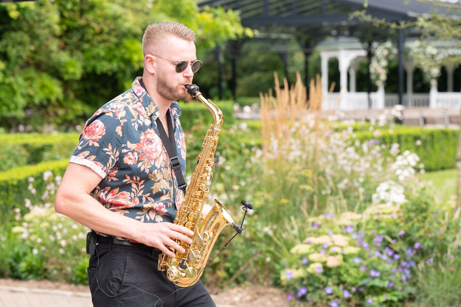 Saxophone player performs in a lush garden setting with blooming flowers and greenery in the background.