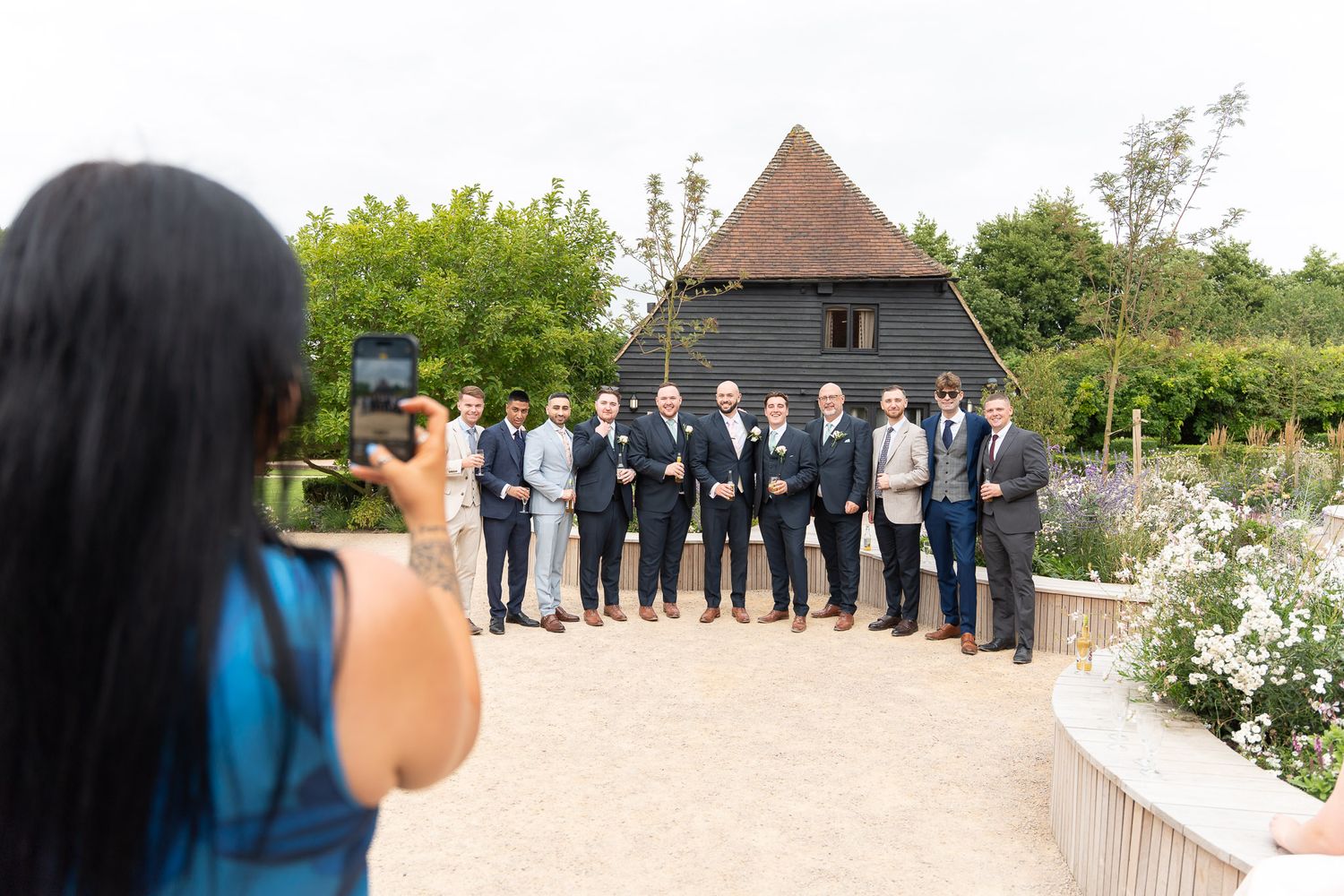 A photographer in a blue shirt takes a group photo on a garden path with white flowers and a gazebo in the background.