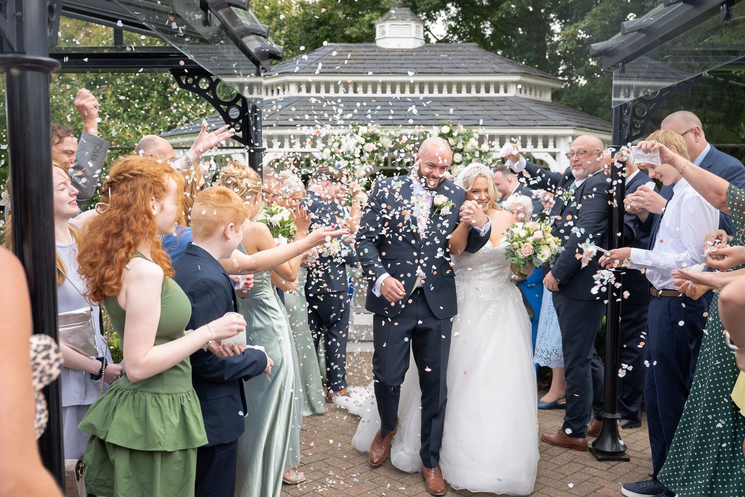 Wedding guests shower newlyweds with confetti during outdoor celebration while bridesmaids in sage green dresses look on.