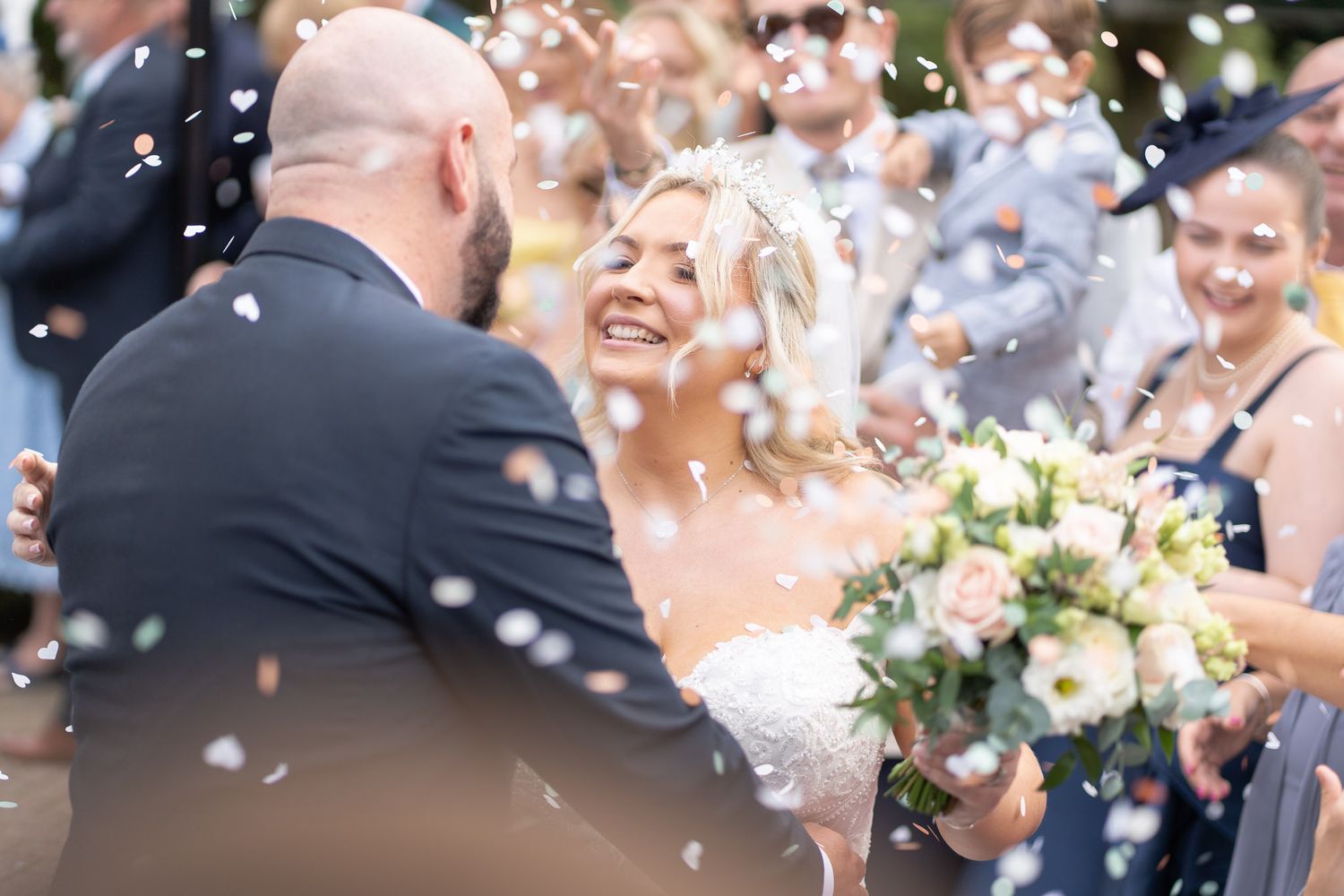 Wedding guests celebrate with confetti as newlyweds share a joyful moment surrounded by white flower bouquets.
