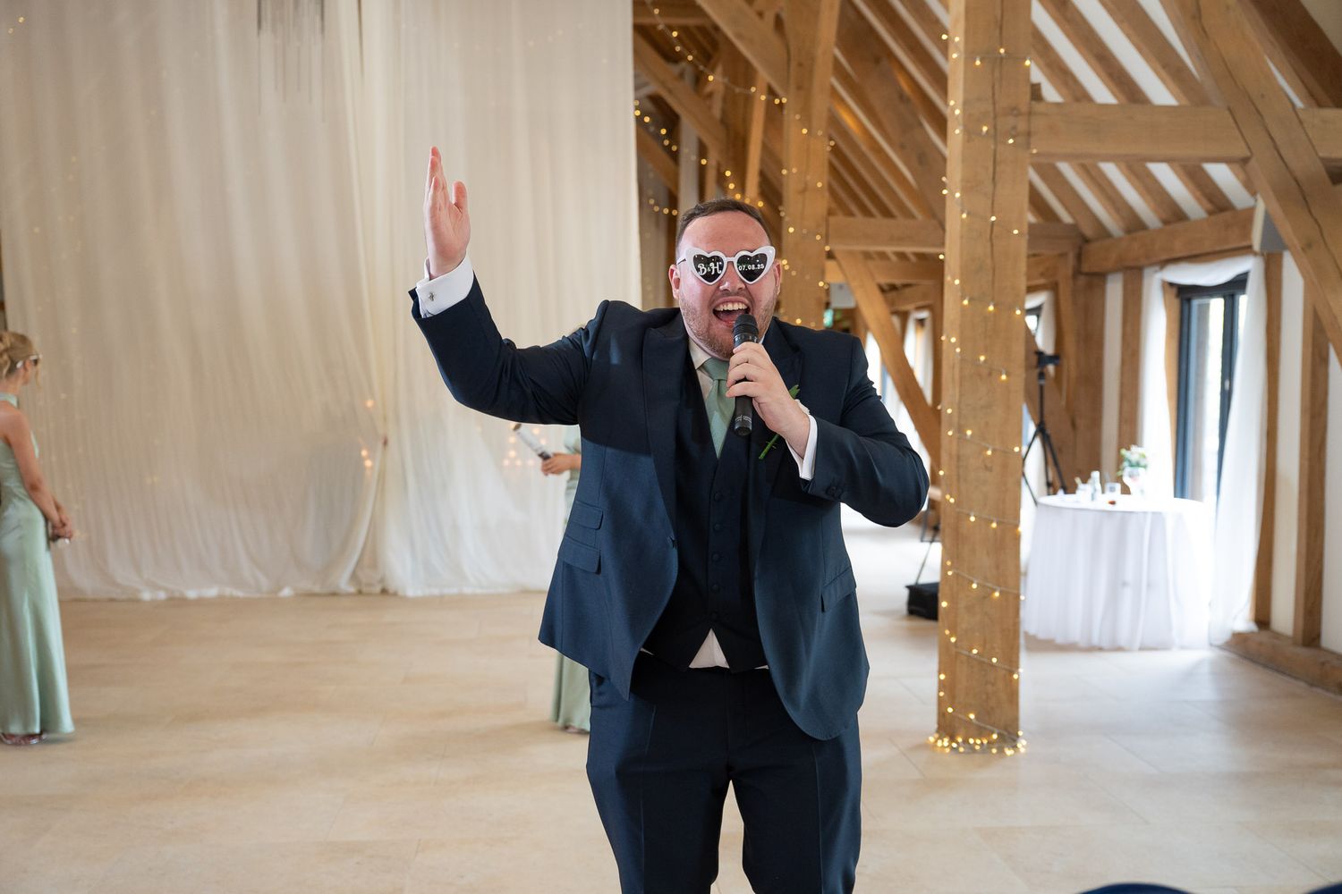 A person in a dark suit gestures enthusiastically in a rustic wooden attic space with exposed beams and natural light.