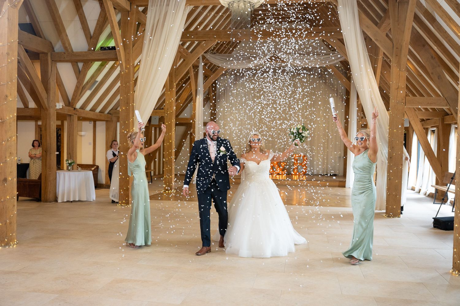 Wedding party celebrates with confetti toss in rustic wooden barn venue with exposed beams and natural lighting.