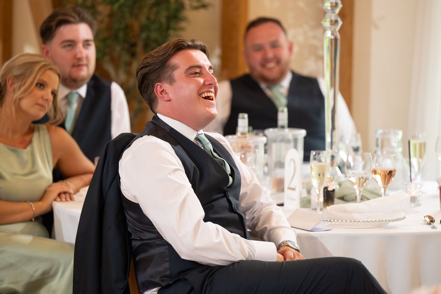 Wedding guests laugh and enjoy themselves at an elegant reception dinner with wine glasses and table settings.