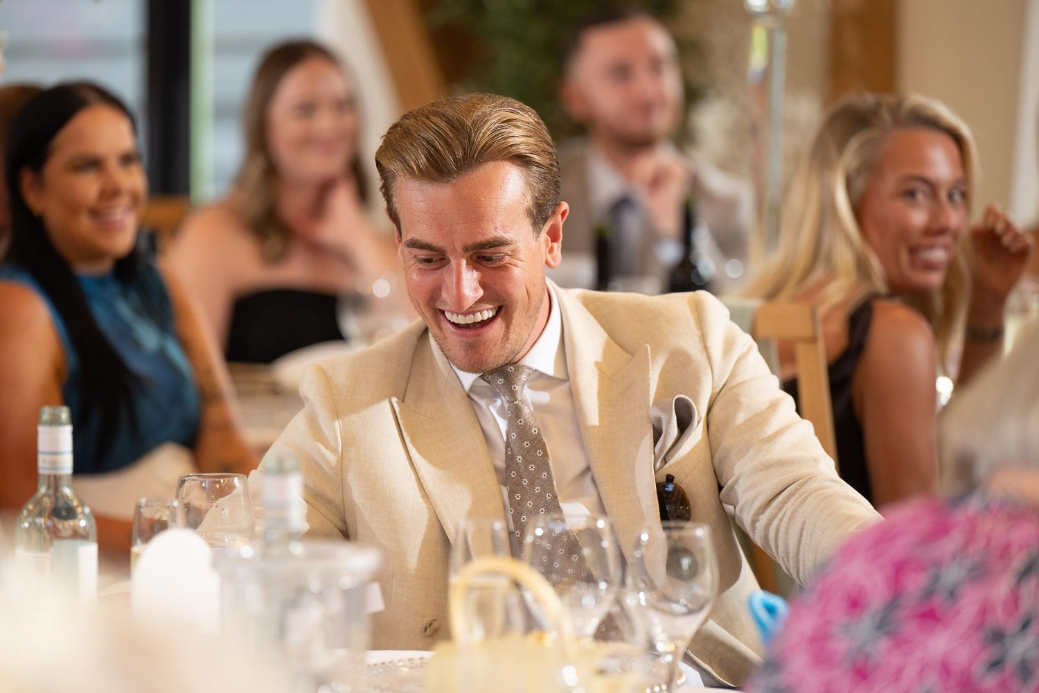 A guest in a cream-colored suit laughs heartily at a formal dinner party surrounded by other attendees.