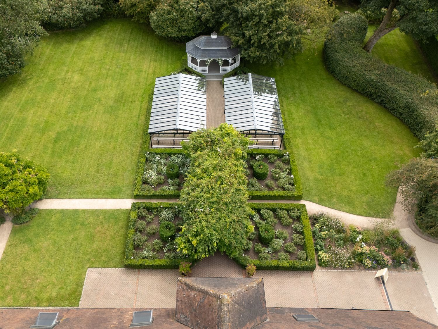 Aerial view of a well-manicured garden with lush green lawn, hedges, and pathways surrounded by trees.