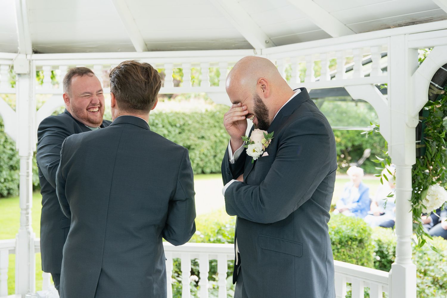A group wearing dark suits share emotional embraces and reactions during an outdoor celebration on a white porch.