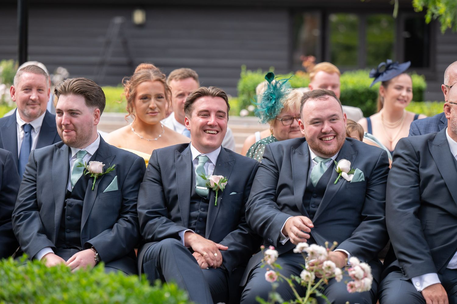 Groomsmen in black suits share a candid laughing moment during an outdoor wedding celebration.