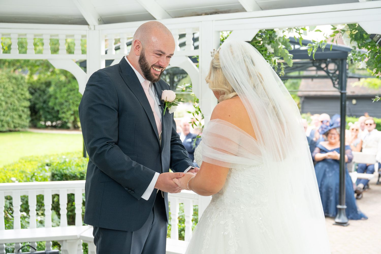 Romantic wedding couple exchanging rings under a white gazebo surrounded by lush greenery.