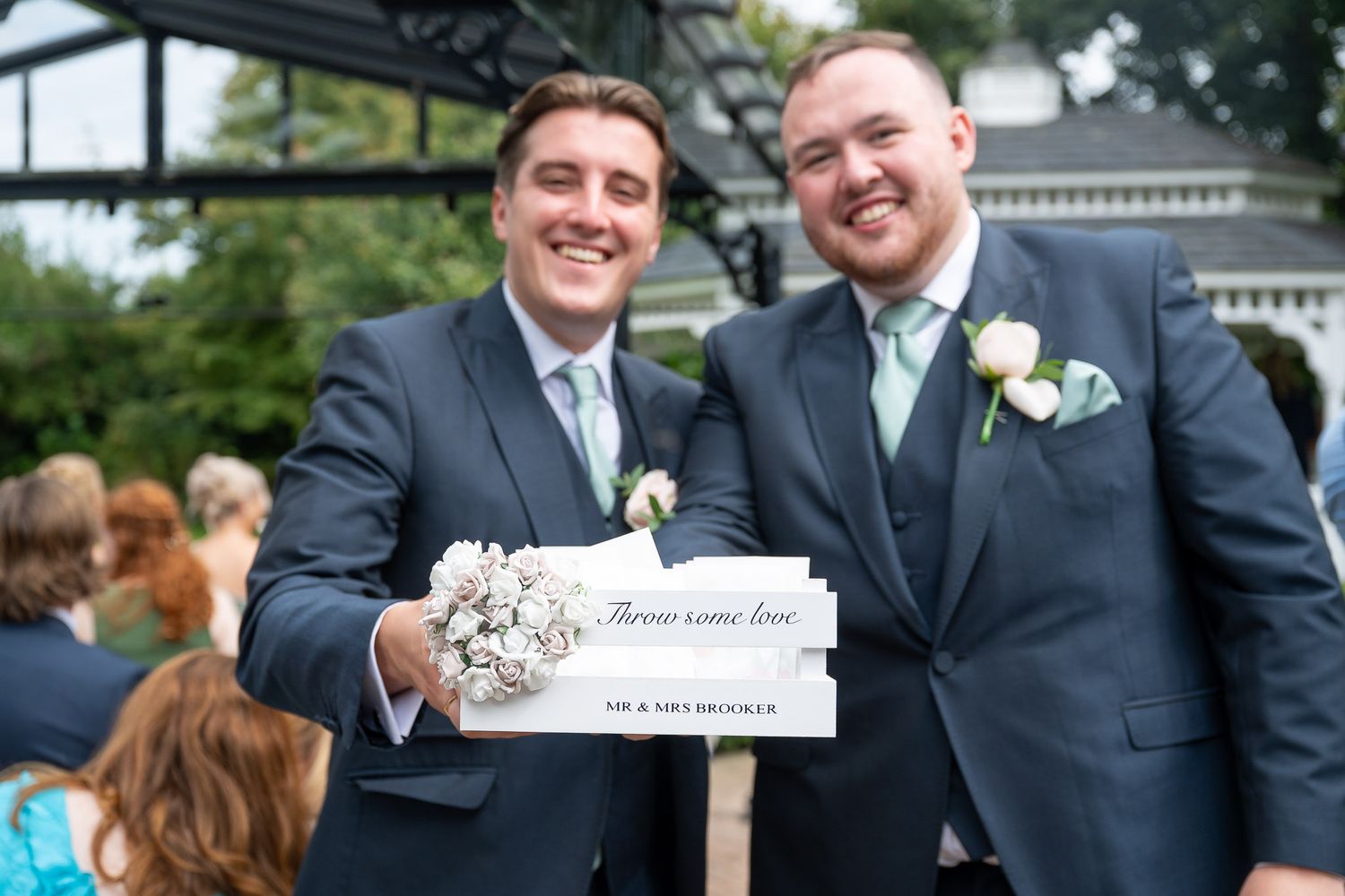 Two people in formal black suits laugh and smile together during an outdoor celebration event.