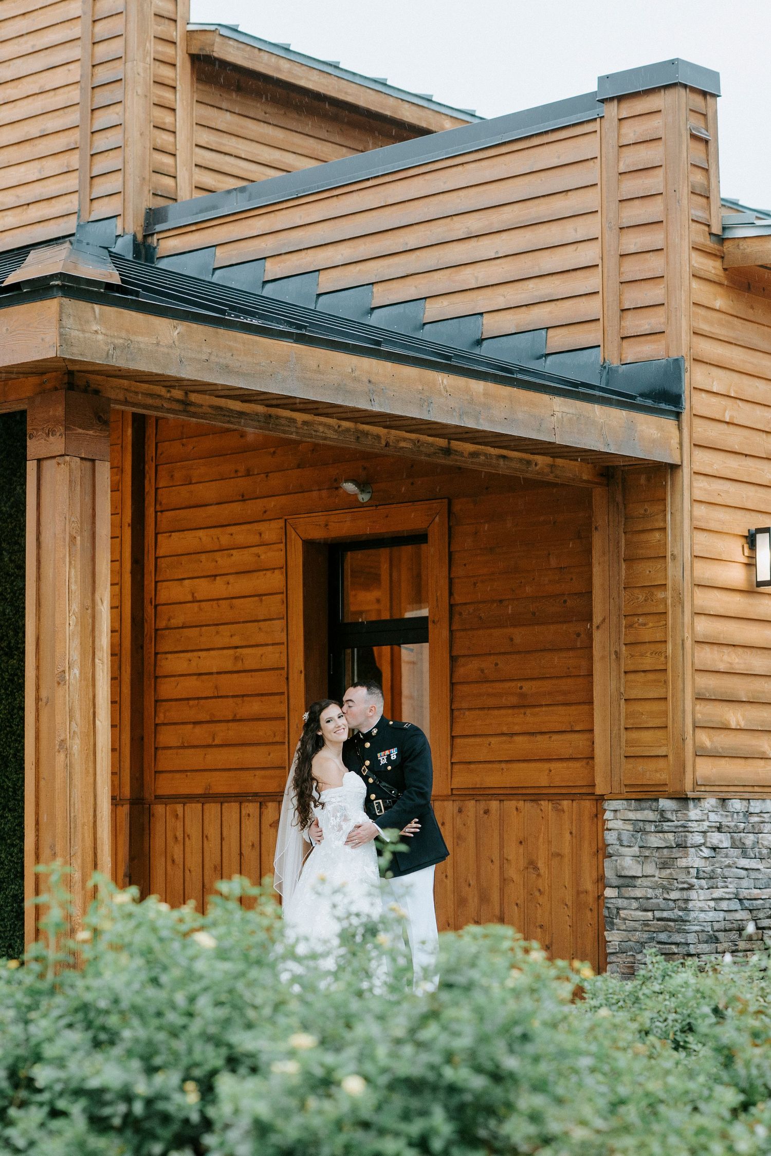 A couple shares an intimate moment under a rustic wooden porch with stone accents and green foliage in the foreground.
