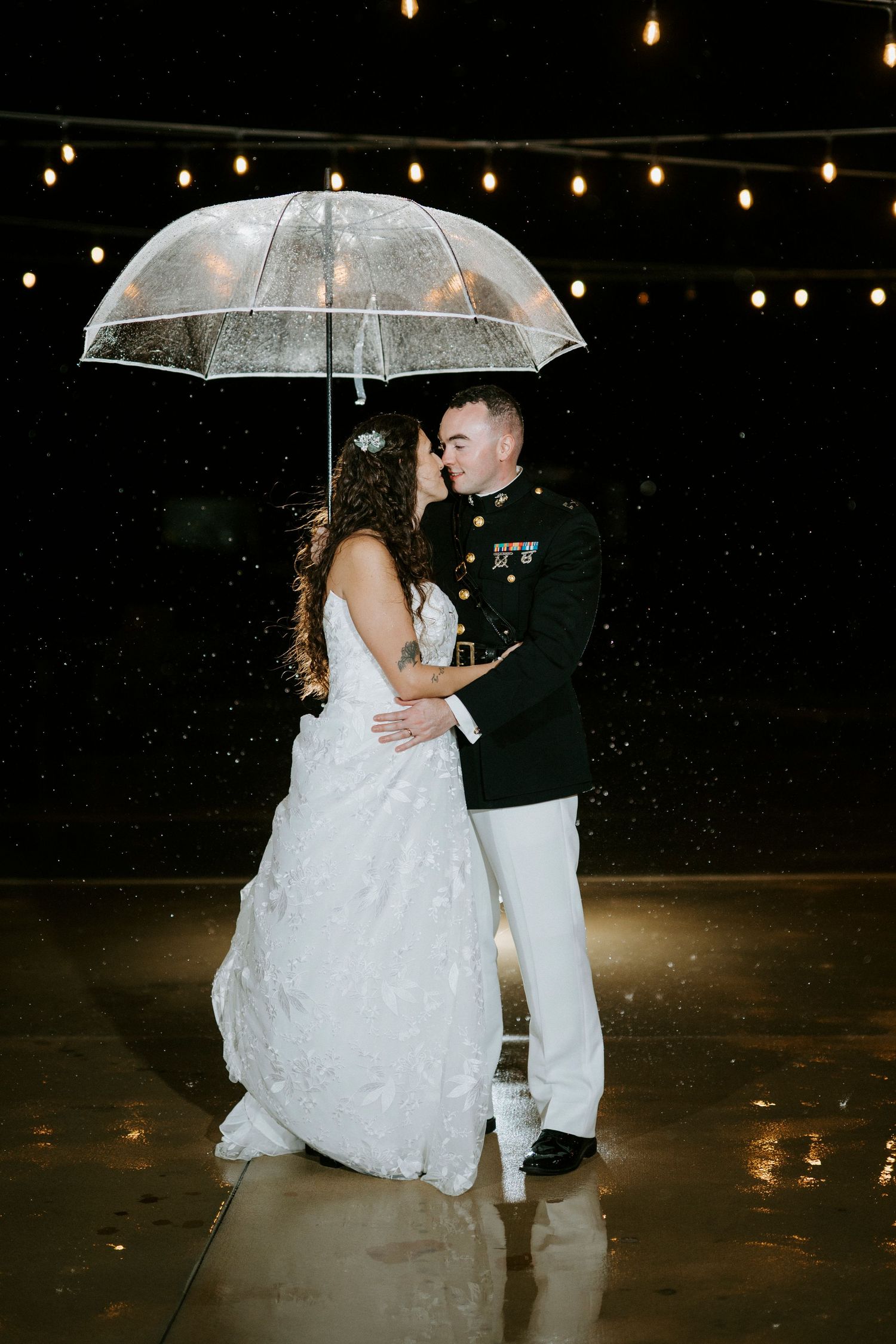 A couple shares a romantic moment under a clear umbrella in the rain with string lights glowing above.
