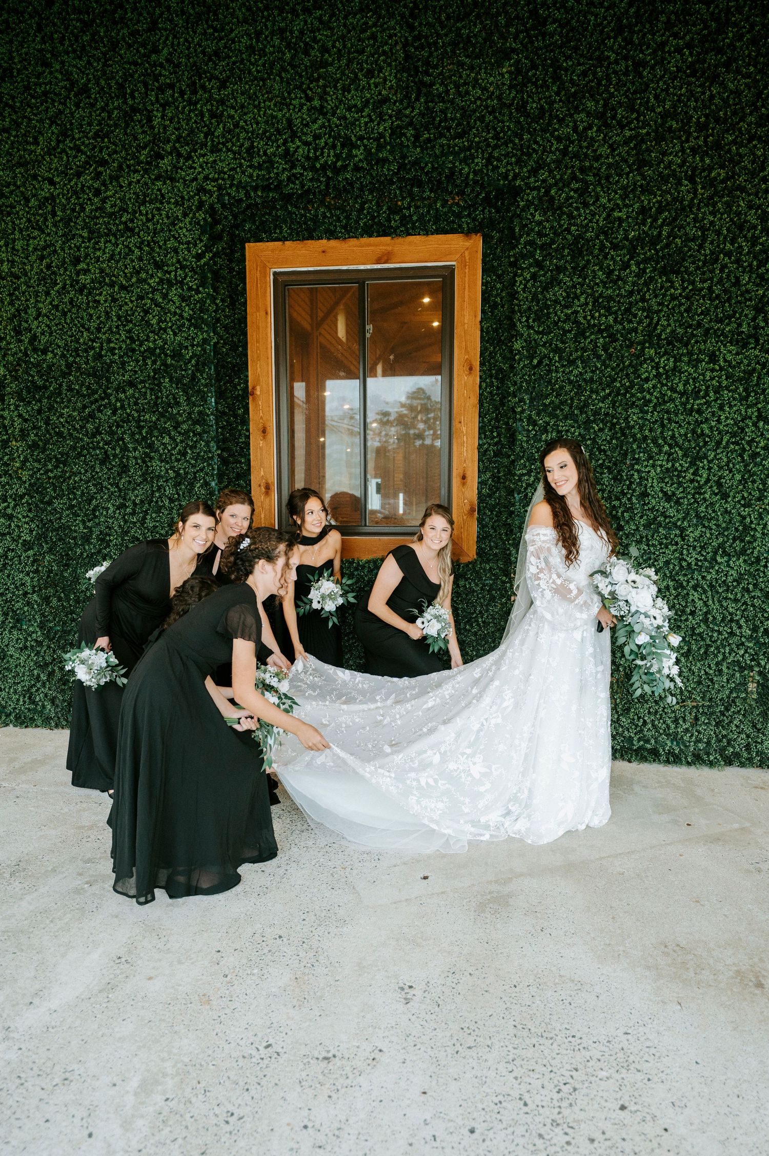 Bridesmaids in black dresses help arrange a long white lace wedding dress train against a green hedge wall backdrop.