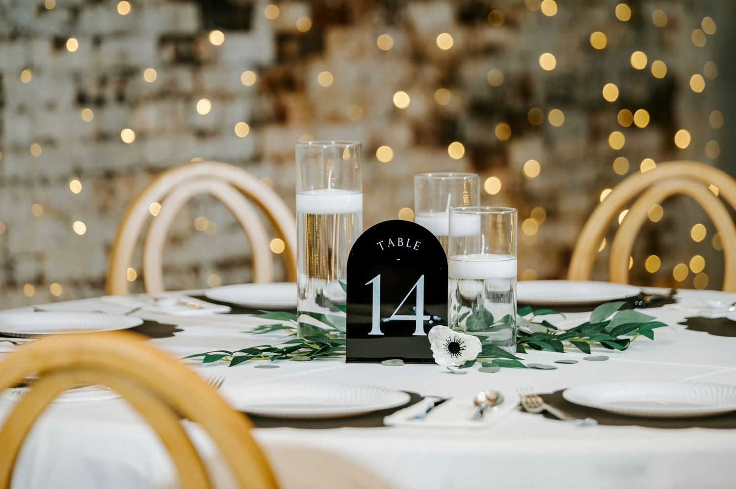 Elegant wedding reception table setting with gold chargers, greenery and twinkling lights in background.