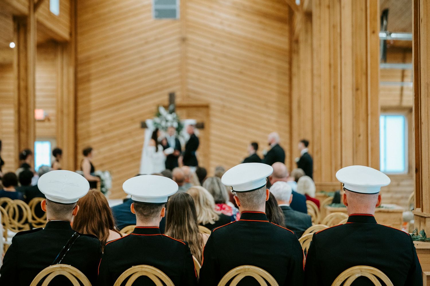 Military service members in dress uniforms sit in rows during a chapel ceremony with wooden walls and natural lighting.