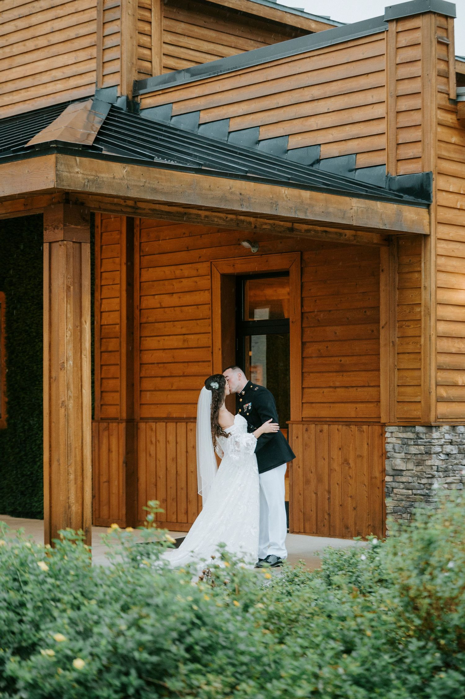 A romantic wedding moment captured outside a rustic wooden lodge with stone accents and greenery.