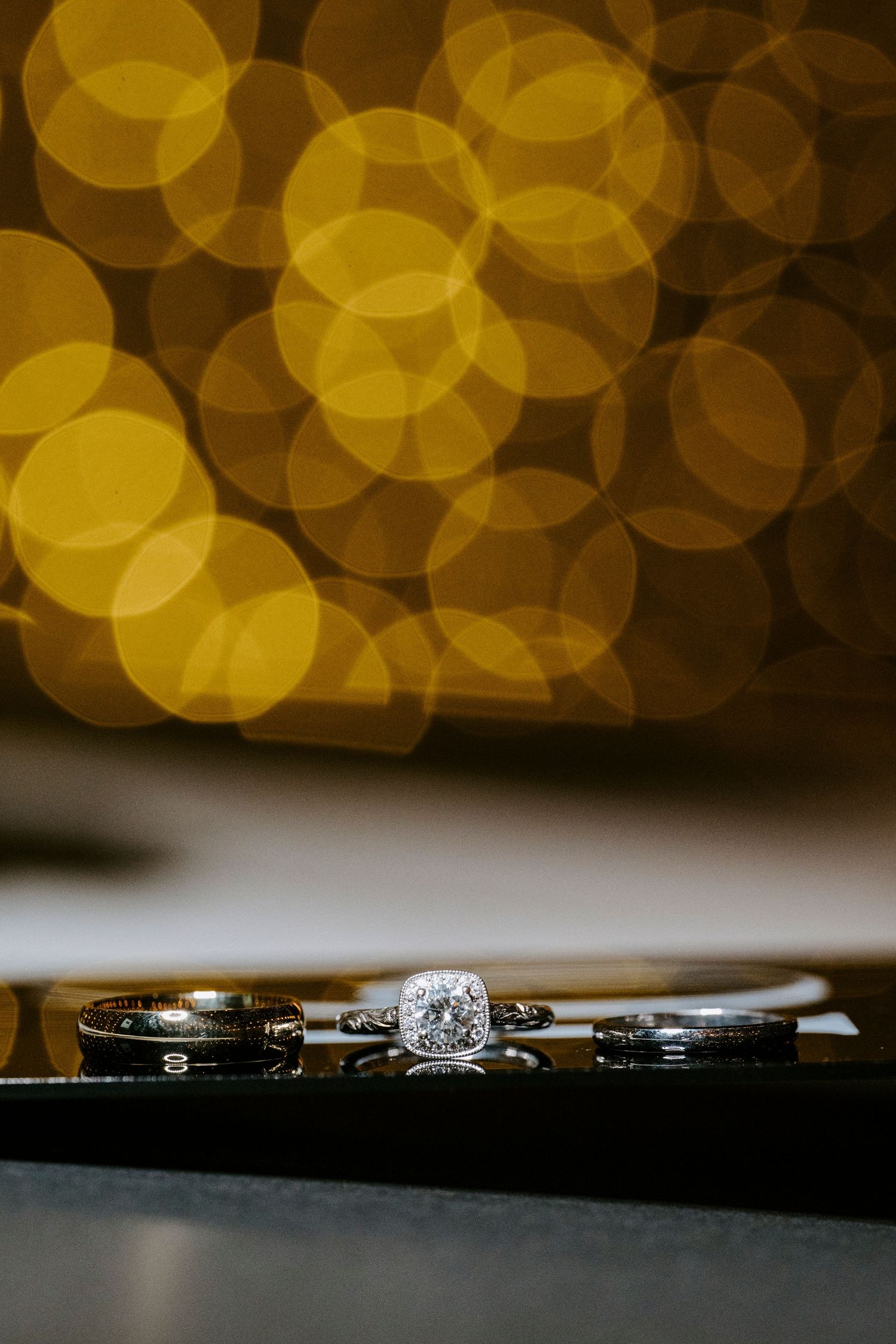 Wedding rings displayed on dark surface with golden bokeh lights creating romantic ambiance in background.