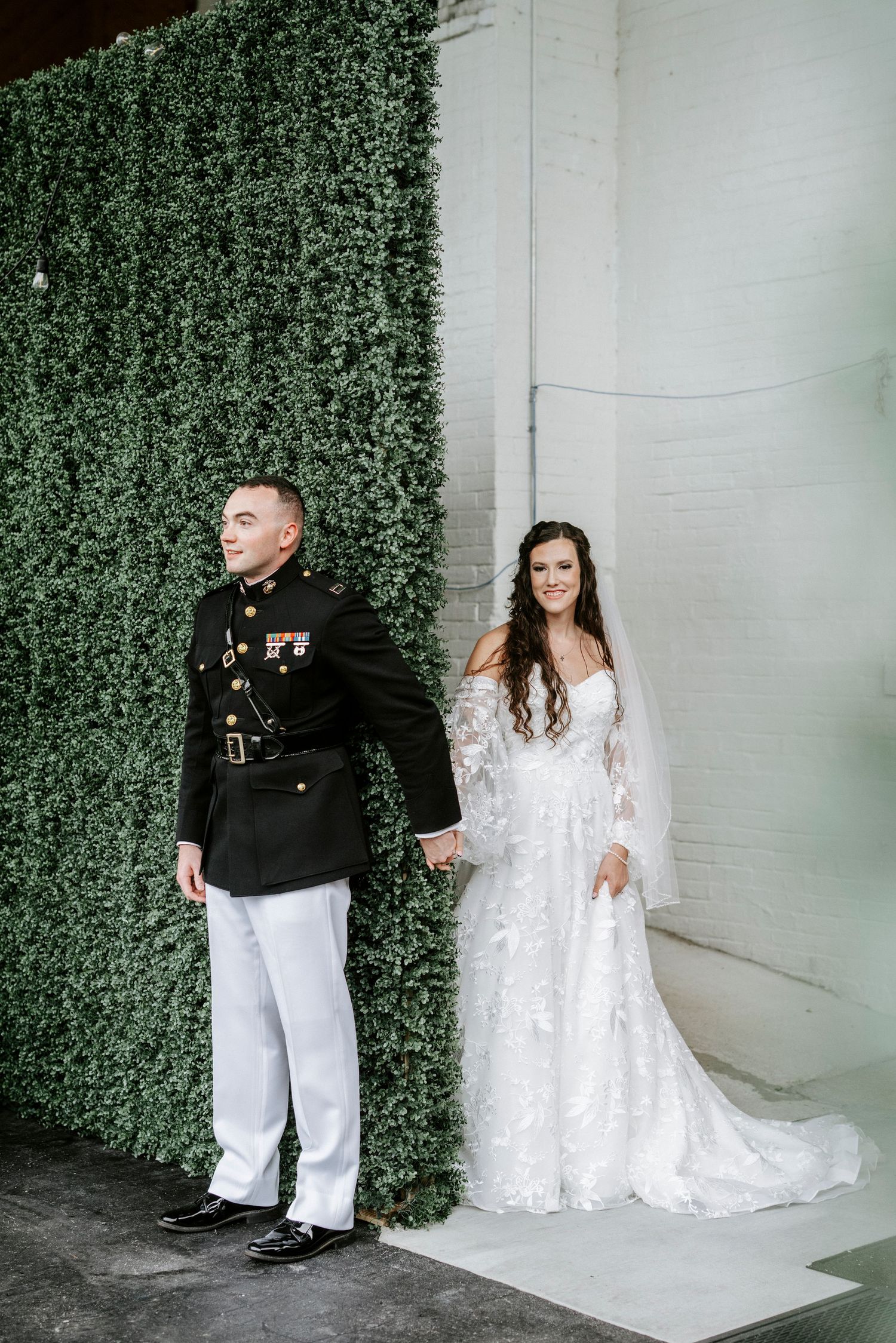 A military groom and bride in wedding attire pose against a green hedge wall for formal portraits.