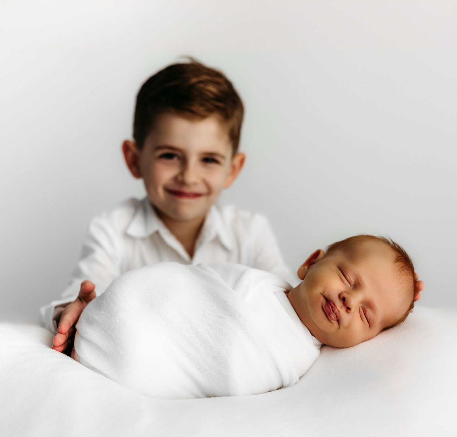A newborn baby lies on white bedding while a child in a white dress leans over to give them a gentle kiss.