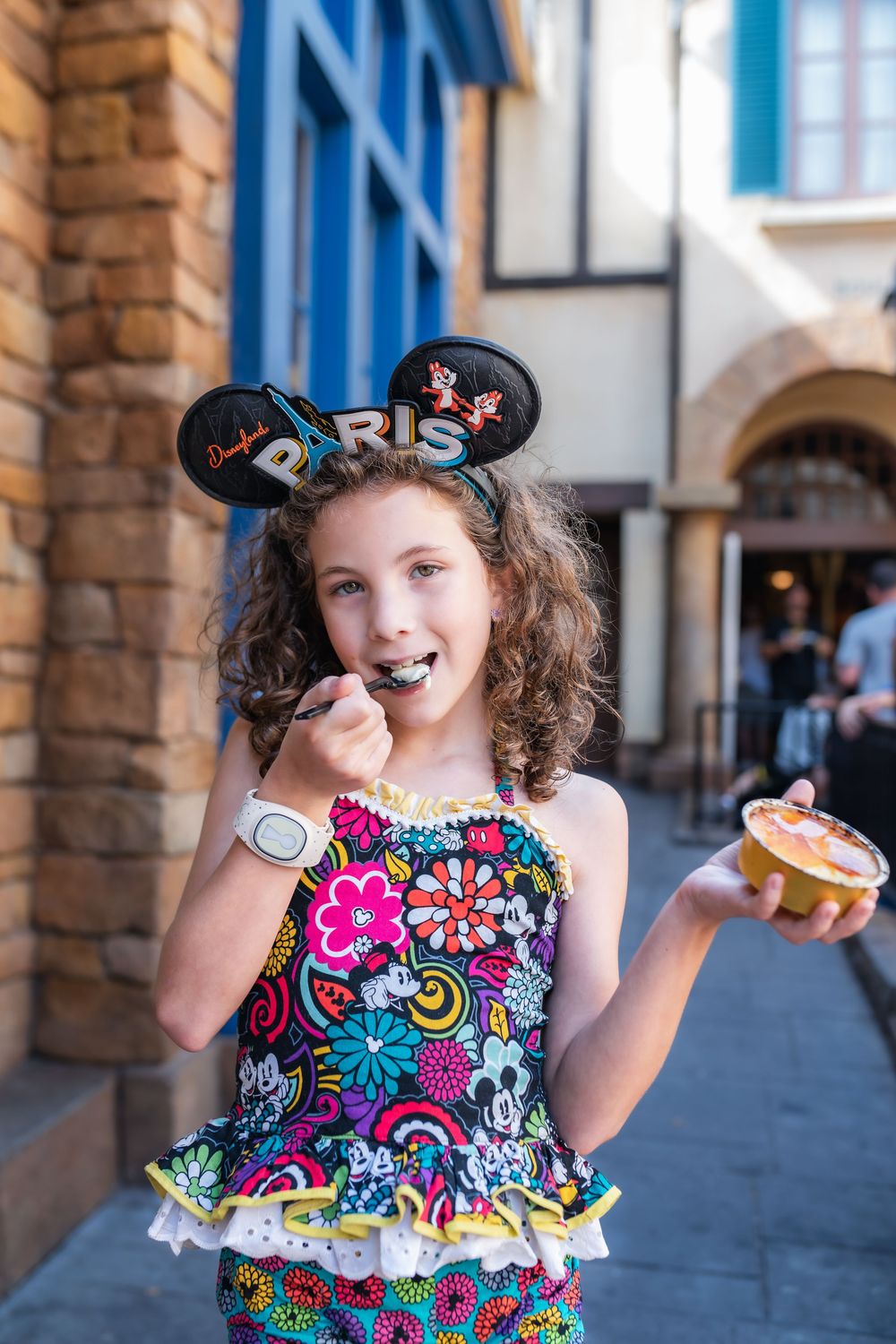 A child in a colorful floral dress poses playfully in front of a stone archway at what appears to be a theme park.