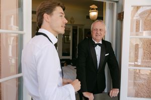 Two people in formal attire having a conversation at a doorway, one in a white shirt and one in a black tuxedo.