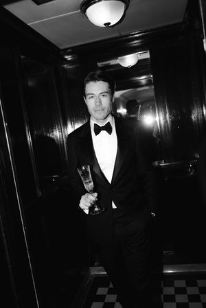 A black and white photo series shows someone in a tuxedo posing in a dimly lit hallway with elegant lighting.