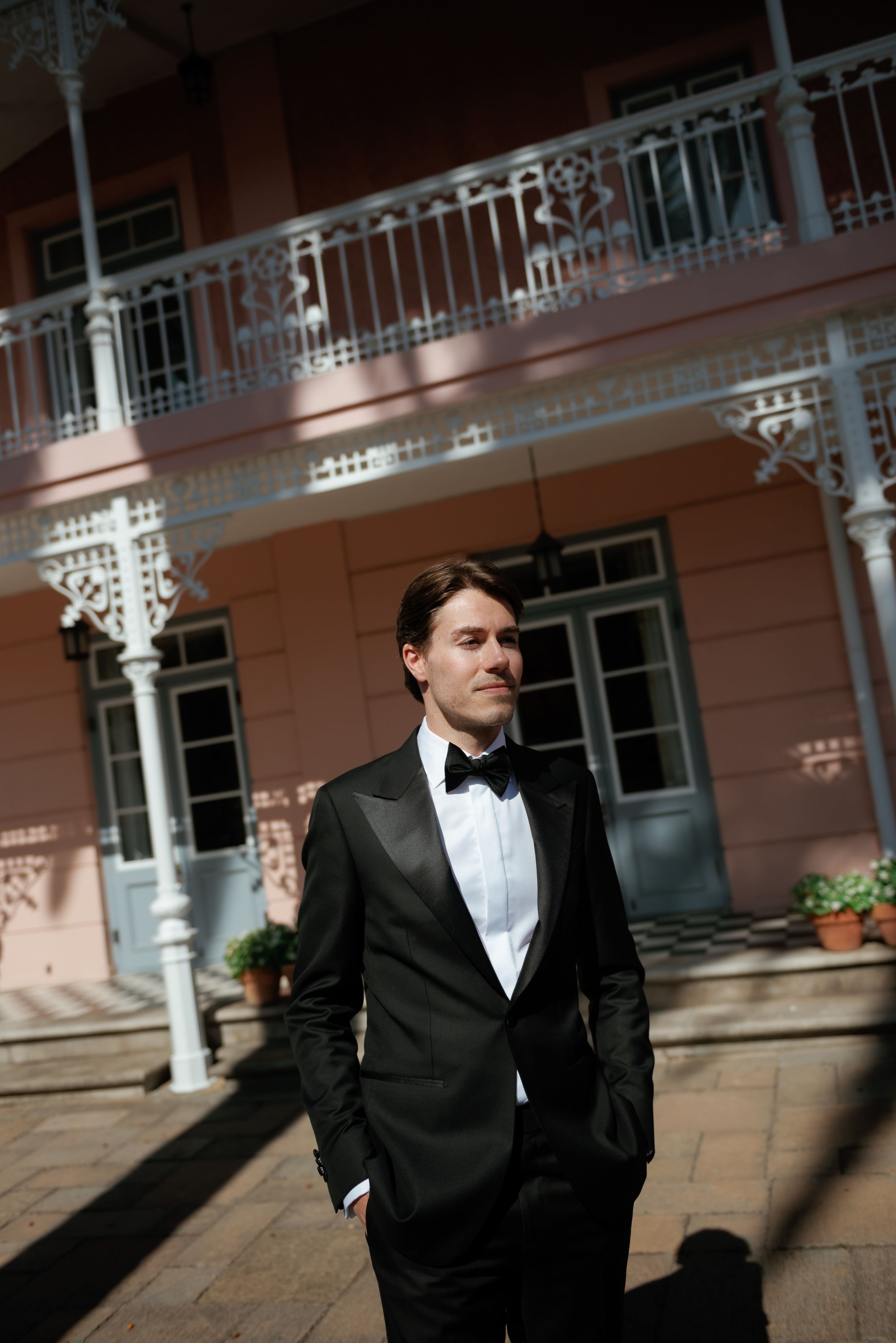 A person in a black tuxedo and bowtie stands against a pink building with ornate white architectural details.
