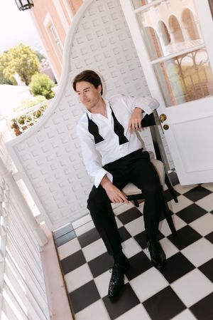 A person in black attire sits on a white chair against a checkered floor and light-colored wall.