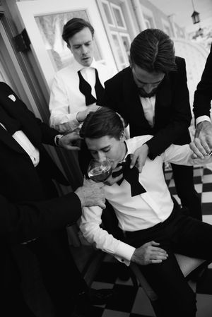 A black and white photo shows groomsmen helping to adjust a bowtie before a formal event on a checkered floor.