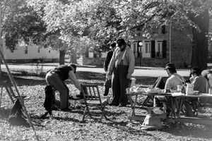 Black and white photo of historical reenactment camp with people gathered under trees.