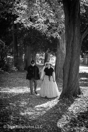Historical reenactors in period clothing walk among tall trees on a leaf-covered path.