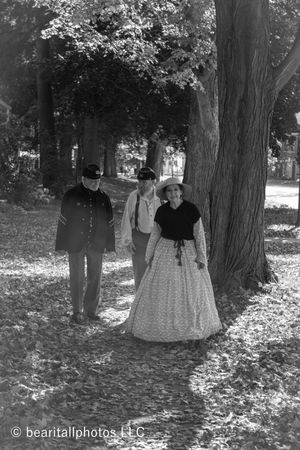 Period-dressed participants walk through a wooded area covered in fallen leaves.