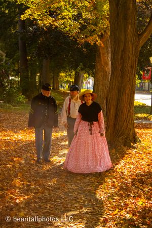 Historical reenactors walk through fallen autumn leaves between large tree trunks.