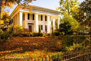 Stately white antebellum mansion with columns surrounded by fall foliage and iron fence.