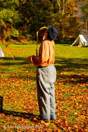 Person in historical clothing walking through fallen autumn leaves with white tents in background.