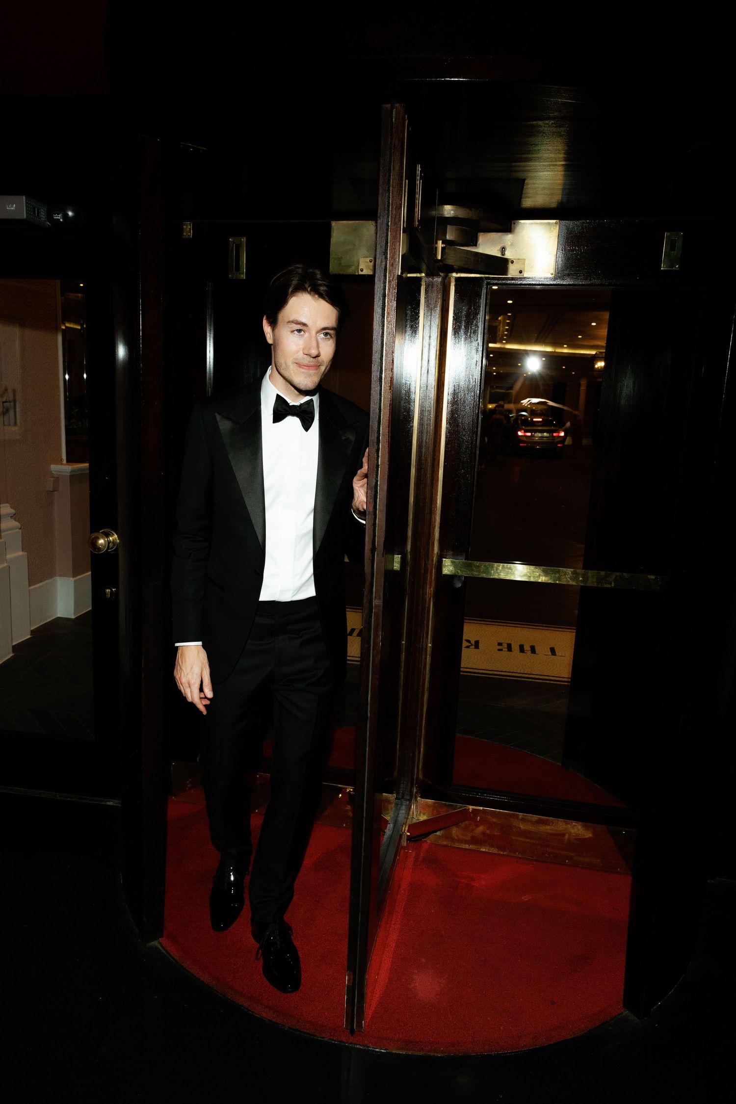 A person in a black tuxedo and bow tie walks on a red carpet at a formal evening event.