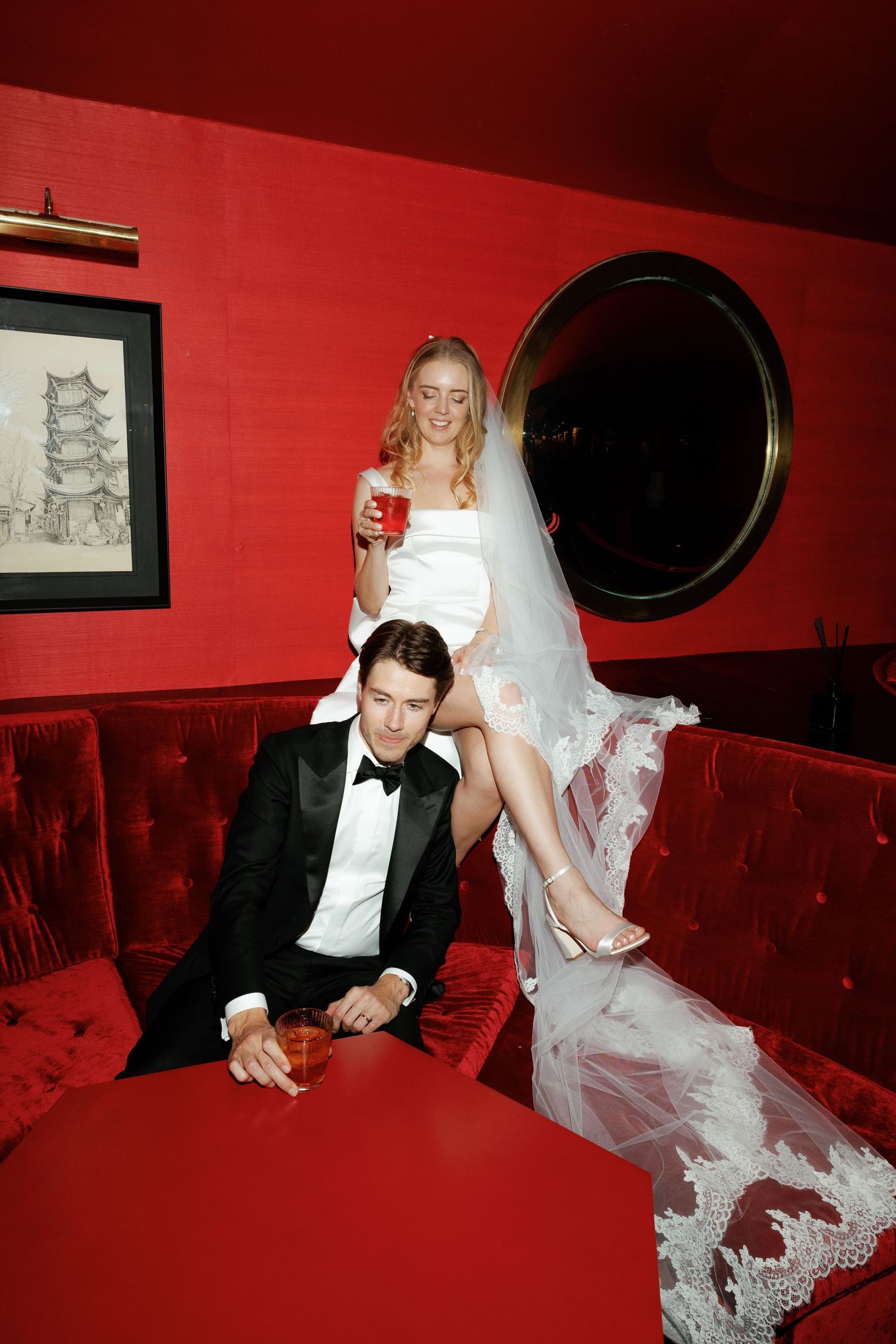 A couple in formal wedding attire posing on a red velvet couch with Asian artwork on the wall.