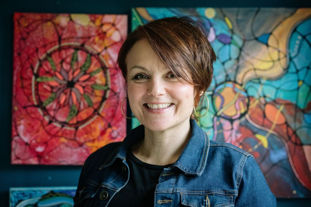 A person in a denim jacket smiles warmly while standing in front of vibrant, colorful abstract wall art.