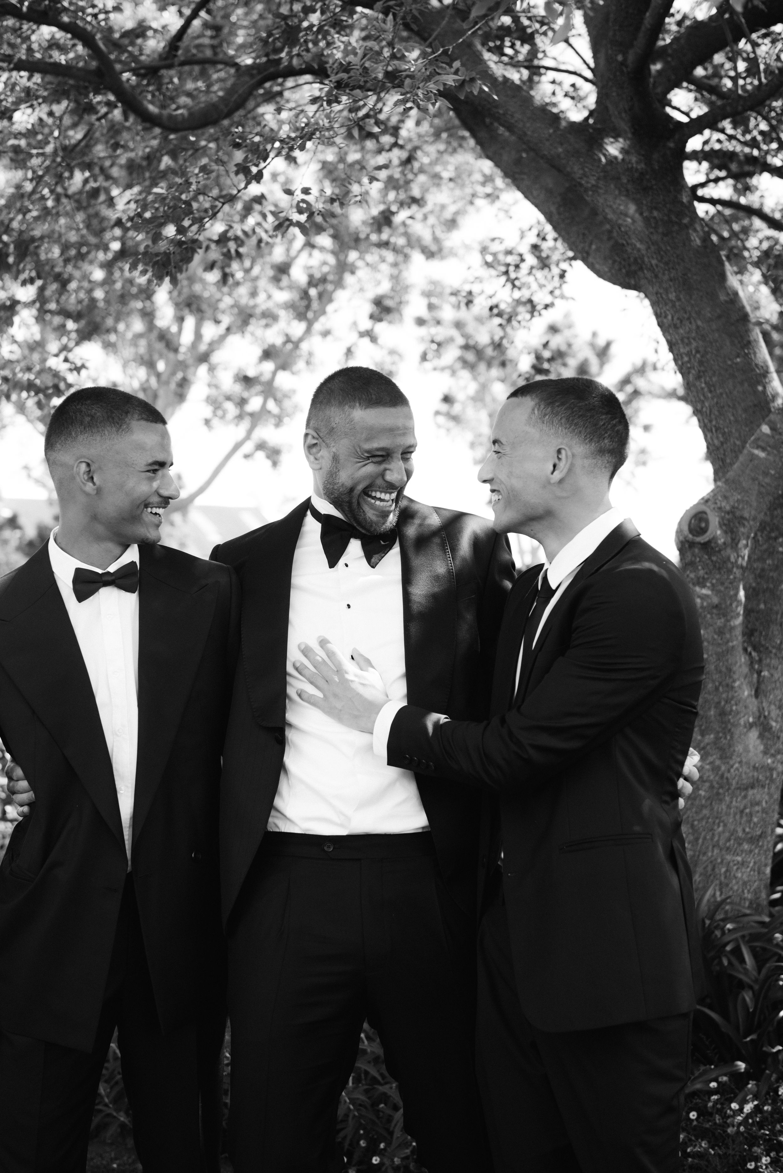 Black and white photo series of groomsmen sharing candid laughing moments under trees at an outdoor wedding.