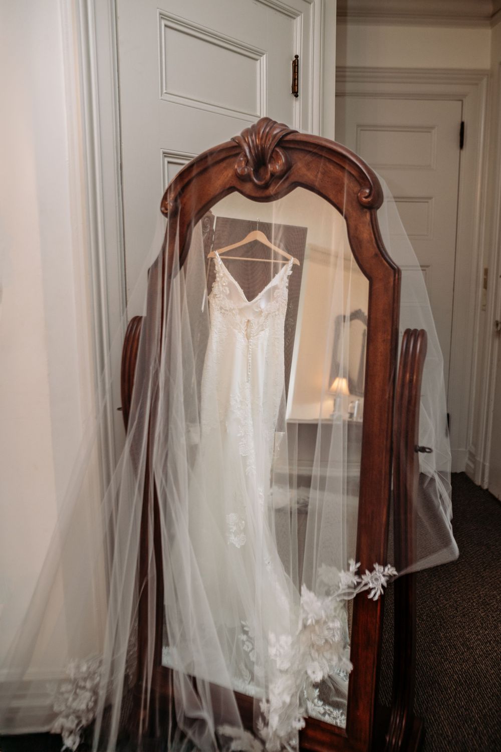A white wedding dress hangs from an ornate wooden mirror frame in a room with white walls.