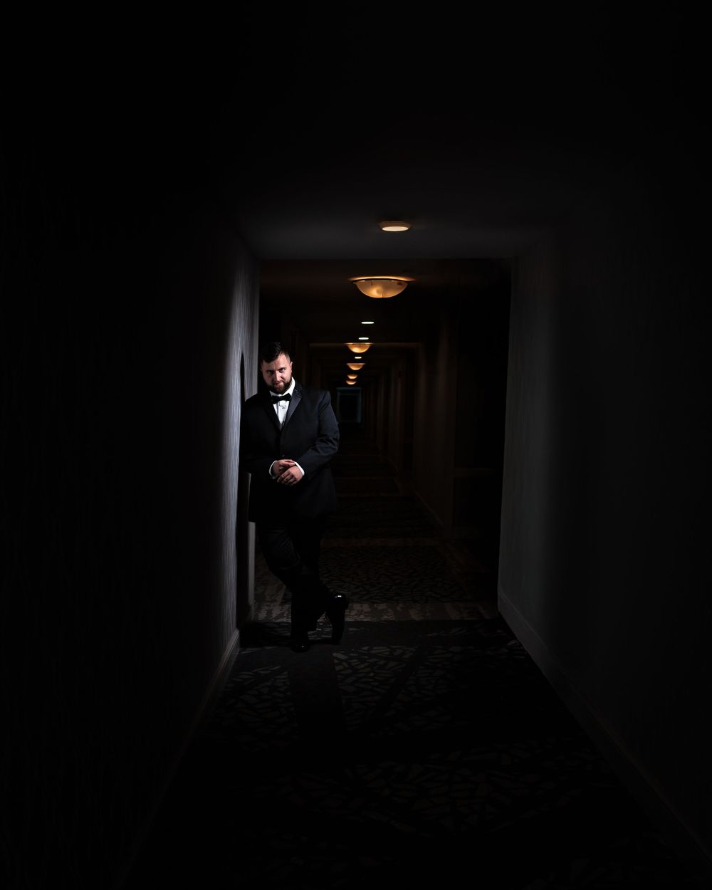 A dimly lit hallway with dramatic lighting shows a figure emerging from darkness in a formal suit.