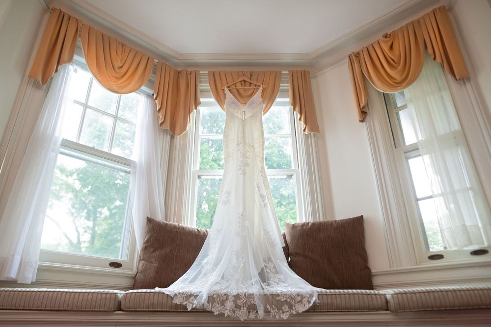 Elegant living room with brown valances and white sheer curtains framing large bay windows overlooking a garden view.
