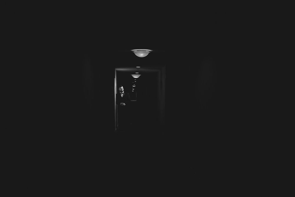 A dark black and white photograph shows a shadowy silhouette standing in a dimly lit space.