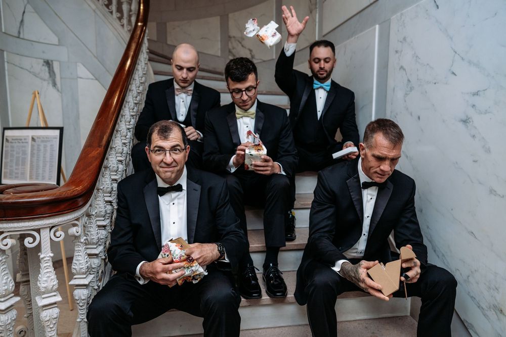 Groomsmen in black tuxedos sit on marble stairs while checking their phones during a formal event.