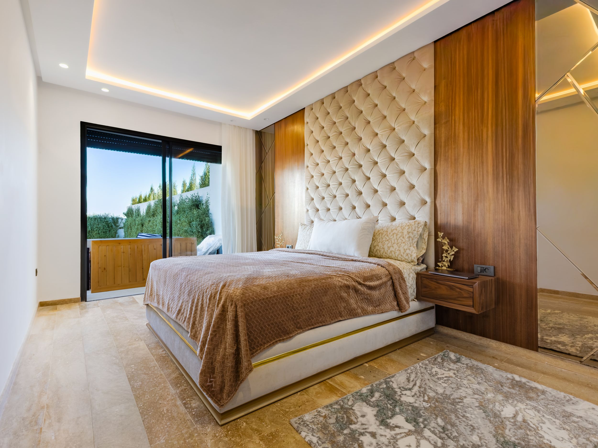 Luxurious master bedroom with tufted gold headboard, wood accent wall, recessed lighting, and balcony access.