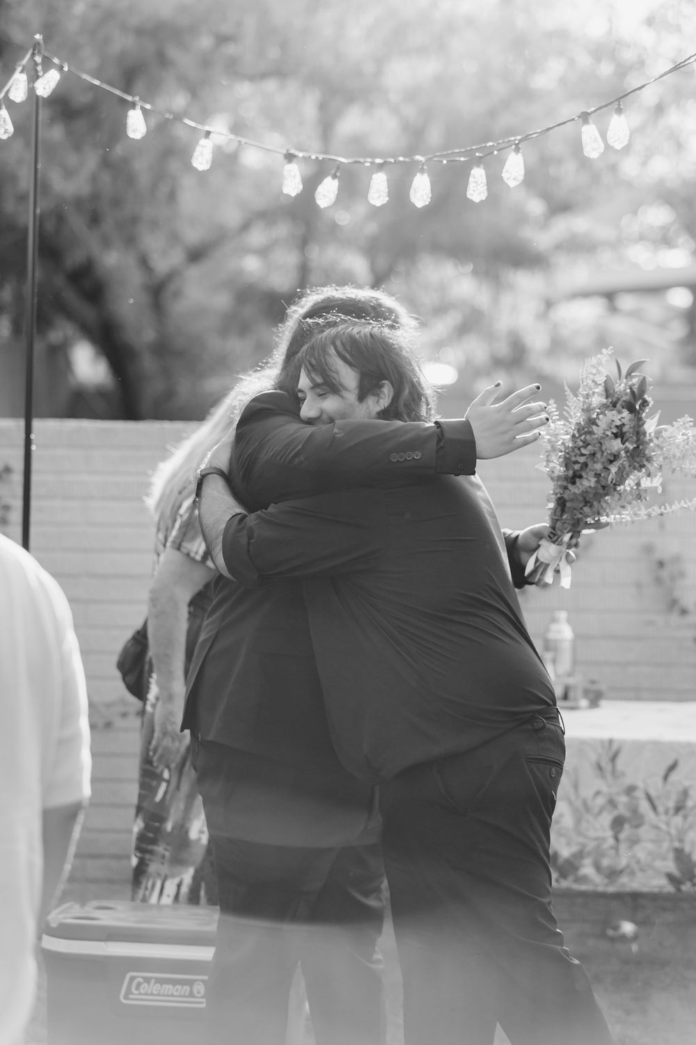 A tender embrace at an outdoor wedding reception with string lights hanging overhead in black and white.