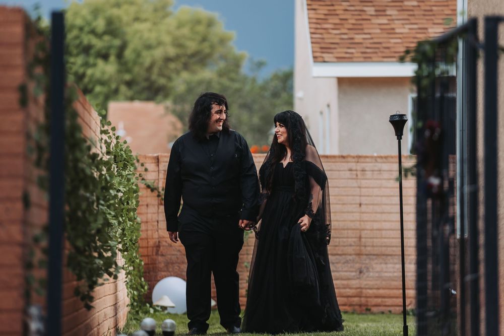 Two people in black clothing walk past brick buildings on a sunny day.